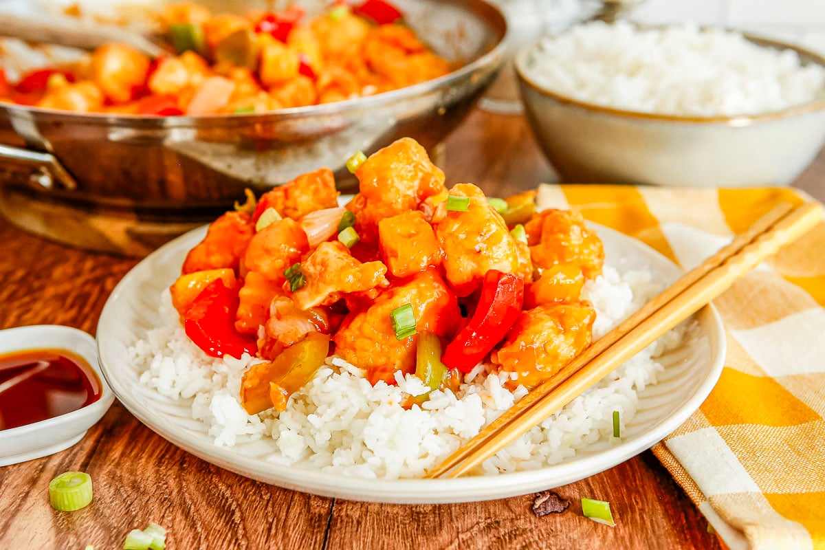 A white plate holding white rice topped with sweet and sour chicken and bell peppers. Wooden chopsticks rest on the edge of the plate next to a small dish of soy sauce.