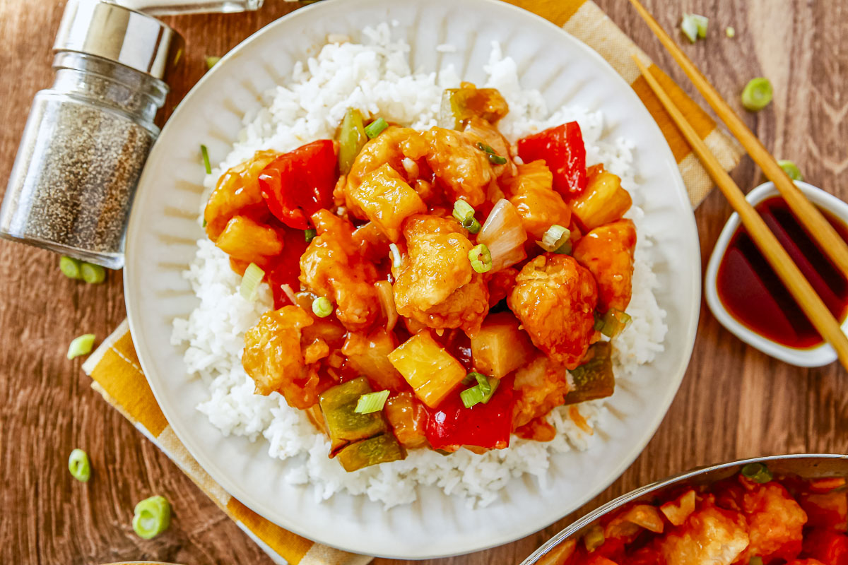 An overhead view of a white plate filled with white rice and homemade sweet and sour chicken. The dish is garnished with green onions and served with chopsticks.