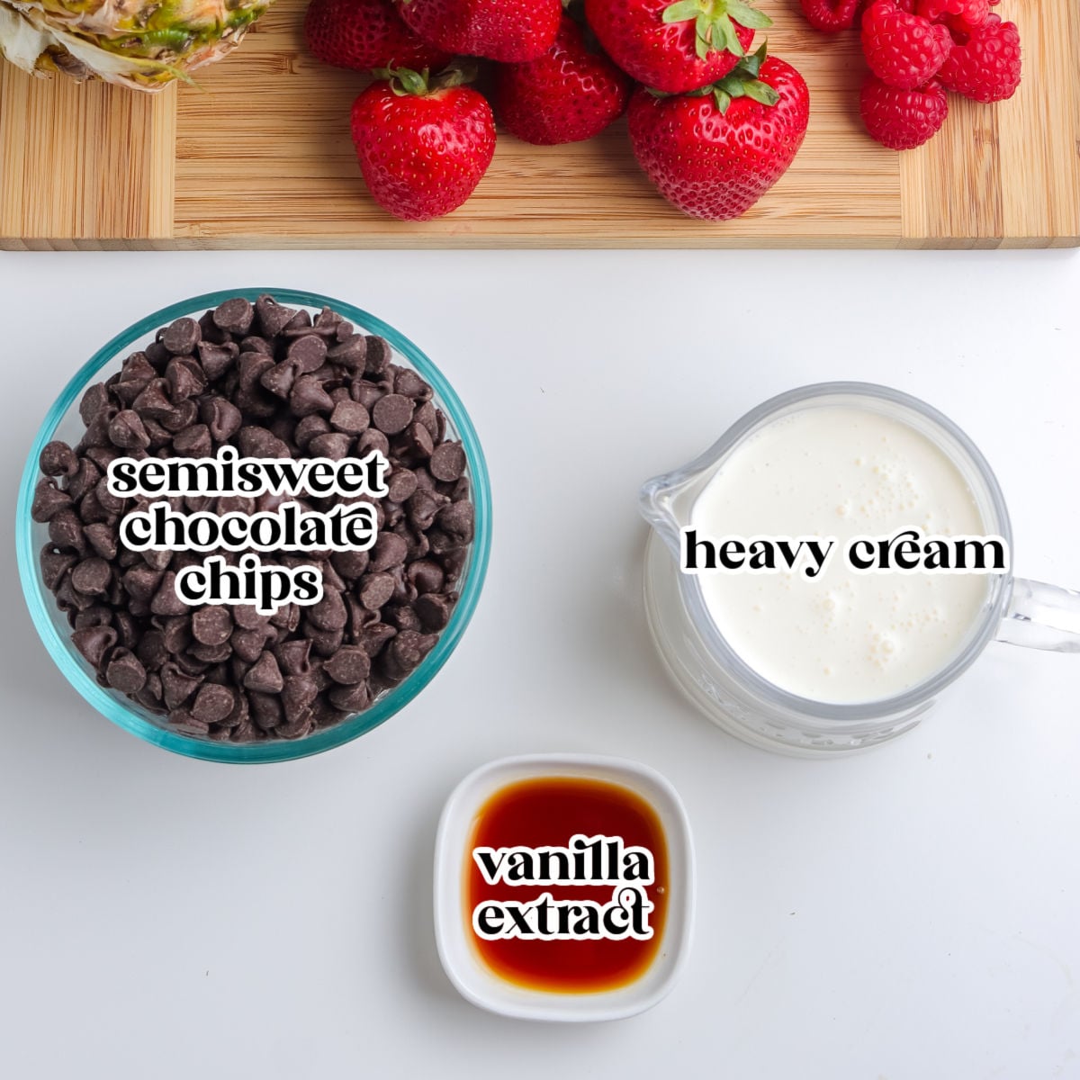 A flat lay showing labeled ingredients needed to make chocolate fruit dip, including a bowl of semisweet chocolate chips, a glass cup of heavy cream, and a small dish of vanilla extract below a wooden board of fresh fruit.