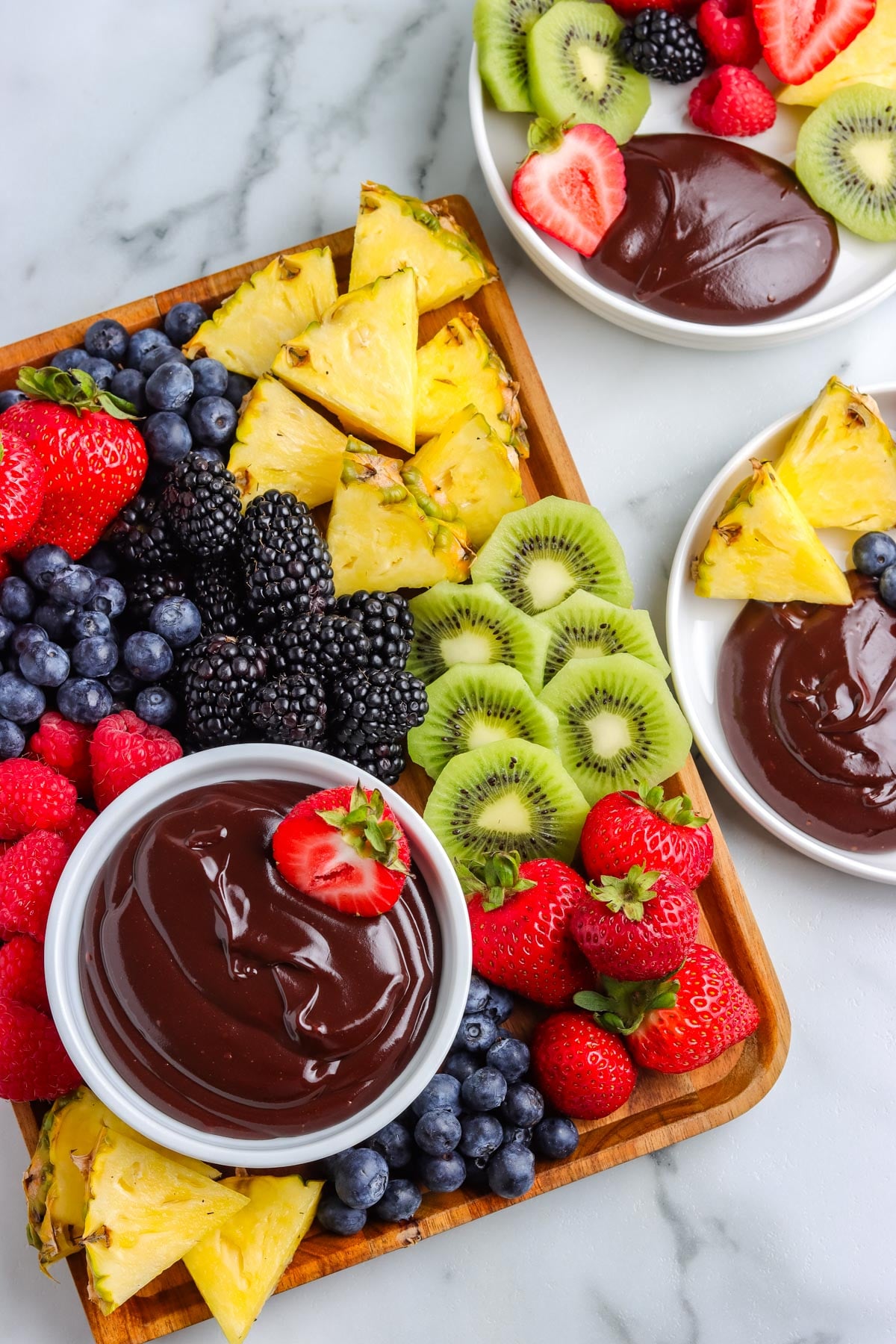 An overhead view of a rectangular wooden dessert board loaded with fresh berries, kiwi slices, and pineapple chunks, featuring a white bowl of chocolate fruit dip in the corner.