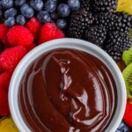 An overhead close-up view of a white bowl filled with glossy chocolate dip, surrounded by fresh blackberries, blueberries, raspberries, strawberries, kiwi slices, and pineapple chunks.
