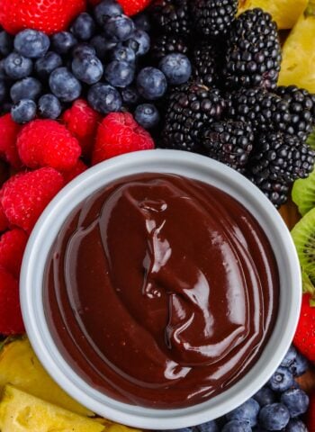 An overhead close-up view of a white bowl filled with glossy chocolate dip, surrounded by fresh blackberries, blueberries, raspberries, strawberries, kiwi slices, and pineapple chunks.
