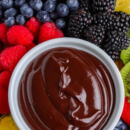 An overhead close-up view of a white bowl filled with glossy chocolate dip, surrounded by fresh blackberries, blueberries, raspberries, strawberries, kiwi slices, and pineapple chunks.
