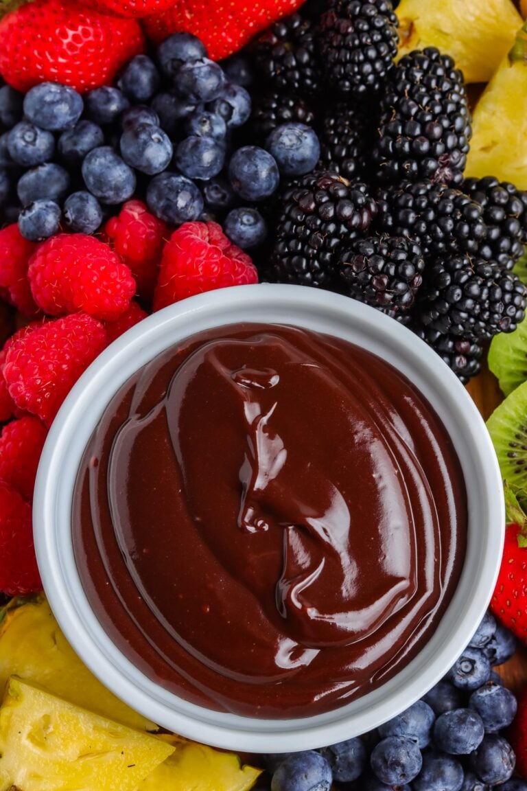 An overhead close-up view of a white bowl filled with glossy chocolate dip, surrounded by fresh blackberries, blueberries, raspberries, strawberries, kiwi slices, and pineapple chunks.
