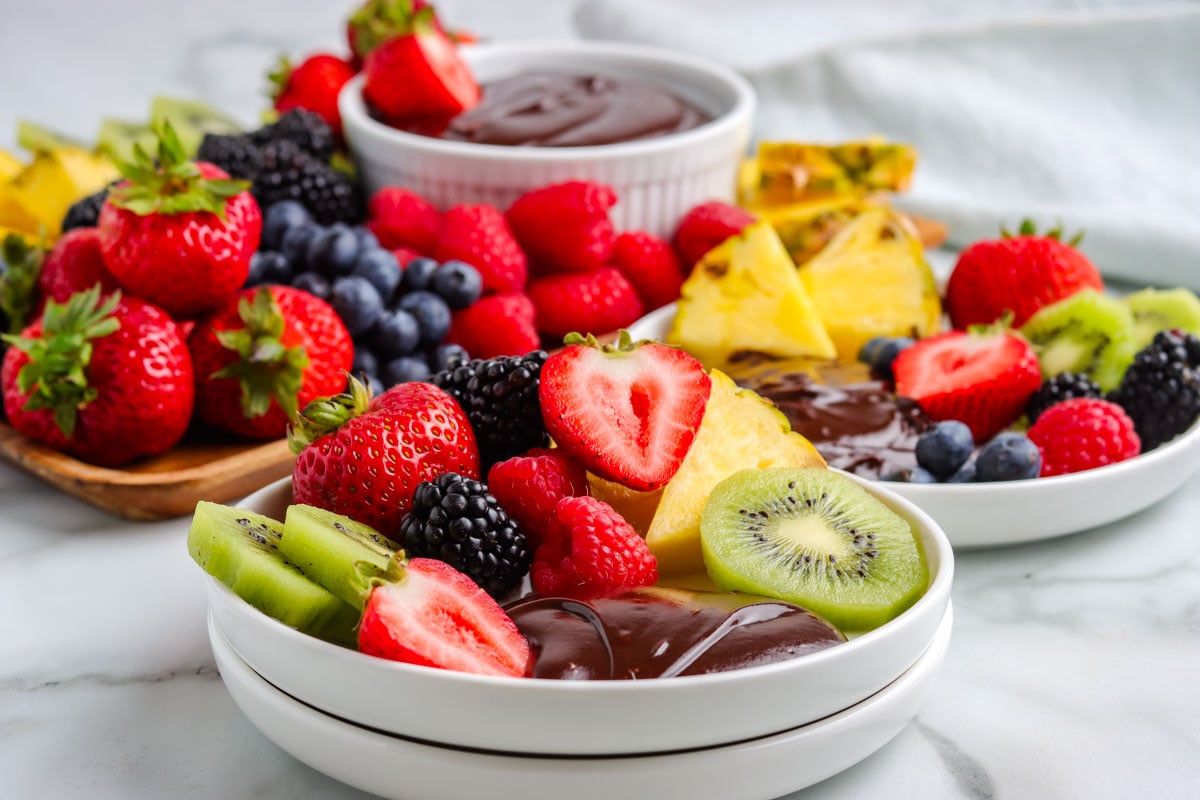 A close-up view of a white plate holding chocolate dip, a sliced strawberry, and kiwi slices, with a large wooden dessert board full of fresh fruit and another bowl of chocolate dip in the background.